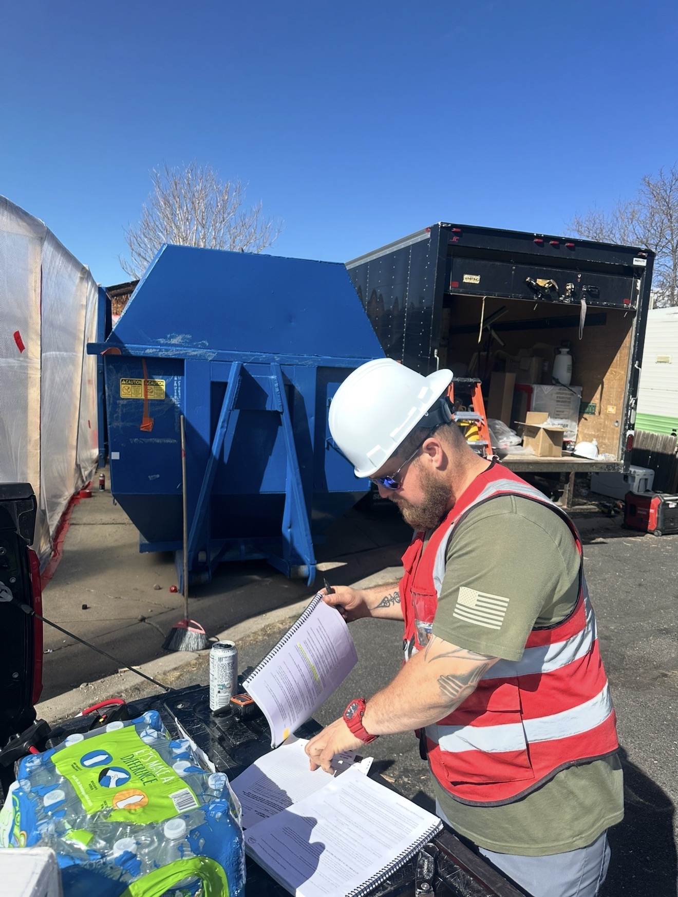 I&F Abatement Experts crew member in hard hat and hi-vis vest reviewing site paperwork next to dump trailer and equipment truck