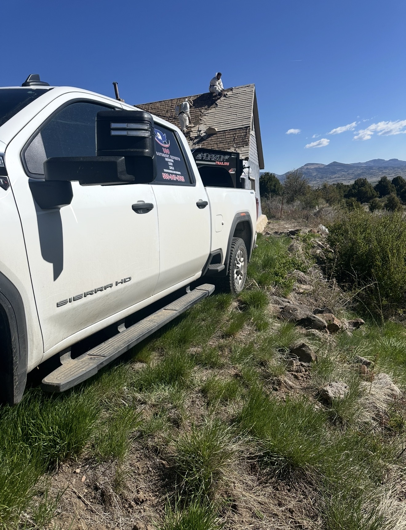 Branded I&F Abatement Experts GMC Sierra HD parked at a remote ridge-top home with crew on the roof during tear-off