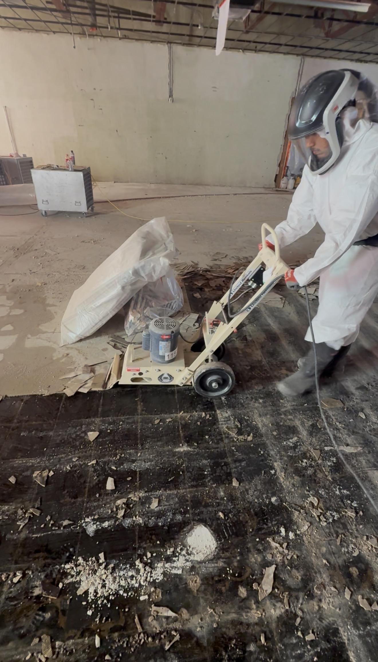 Technician in white Tyvek suit and PAPR helmet operating a floor scraper to remove asbestos floor tile and mastic